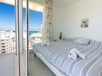 Helles Schlafzimmer mit Doppelbett, gestreifter Bettwäsche, Nachttischlampe und direktem Zugang zum privaten Balkon mit Meer- und Stadtblick.