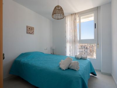 Helles Schlafzimmer mit Doppelbett in Türkis, einer Hängeleuchte aus Naturfasern und einem Fenster mit urbanem Blick auf umliegende Gebäude.