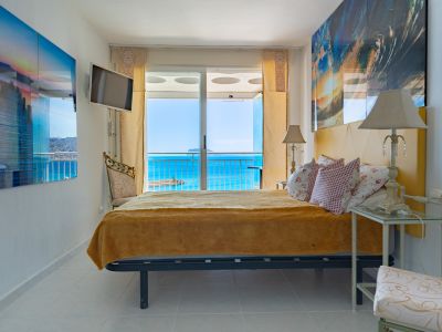 Helles Schlafzimmer mit Doppelbett und gelbem Kopfteil, weißen Fliesen und direktem Zugang zur Terrasse durch Schiebetüren, mit weitem Blick auf das Meer und den Hafen.