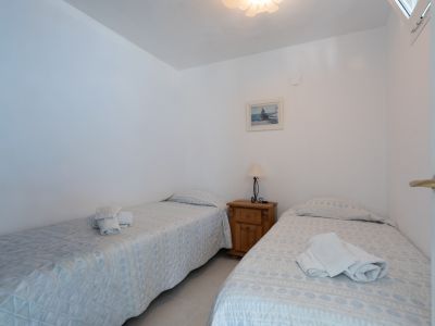 Helles Schlafzimmer mit zwei Einzelbetten, gemusterten Tagesdecken und weißen Handtüchern. Ein Holznachttisch mit Lampe und ein gerahmtes Bild schmücken den Raum.