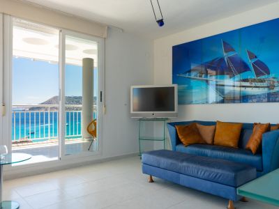 Helles Wohnzimmer mit Glasschiebetüren zur Terrasse und Panoramameerblick. Ausgestattet mit einem blauen Sofa, einem Wandfernseher und einem großen Bootsgemälde.