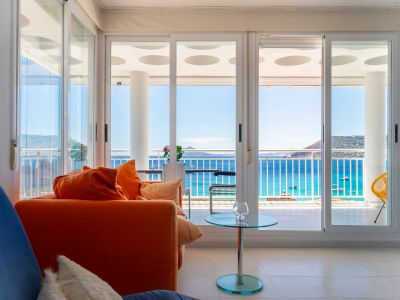 Helles Wohnzimmer mit orangefarbenem Sofa und Glastisch. Große Schiebetüren öffnen zu einer Terrasse mit direktem Meerblick auf Boote und ferne Berge.