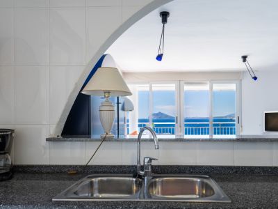 Küchenbereich mit Doppelspüle und dunkler Arbeitsplatte, durch einen Bogen mit dem Wohnbereich verbunden, der eine Terrasse mit weitem Meerblick freigibt.