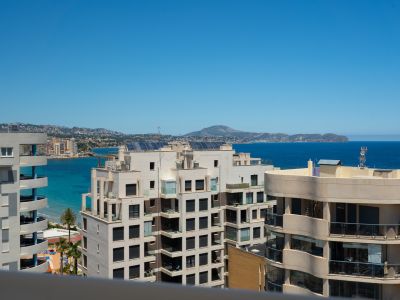 Panoramablick auf das Meer von einer Hochterrasse, mit moderner Küstenarchitektur, türkisfarbenem Wasser und fernen Bergen unter klarem blauem Himmel.