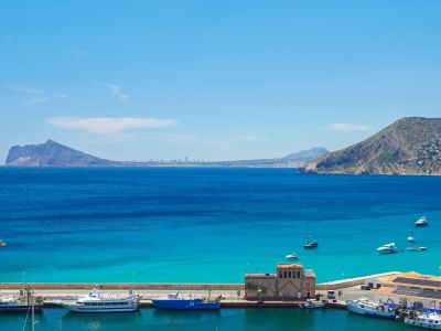 Panoramablick auf den Hafen von Calpe mit Booten, türkisfarbenem Wasser und dem ikonischen Peñón de Ifach im Hintergrund, umgeben von der Küstenbebauung.