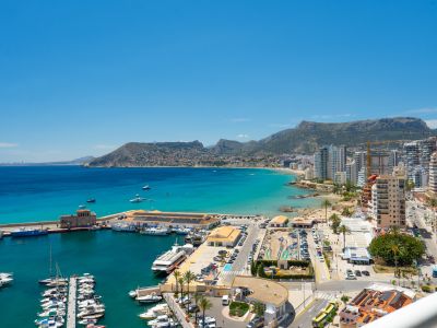 Panoramablick auf die Bucht von Calpe, den geschäftigen Yachthafen, Sandstrände und die Küstenstadt, umrahmt von fernen Bergen unter einem klaren blauen Himmel.