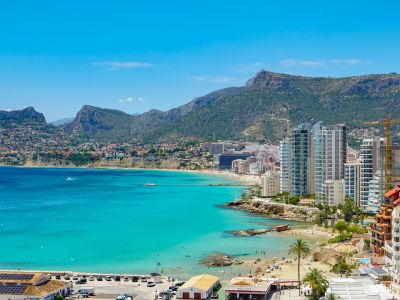 Panoramablick auf die Bucht von Calpe mit türkisfarbenem Wasser, Sandstränden, Küstengebäuden und fernen Bergen unter klarem blauem Himmel.
