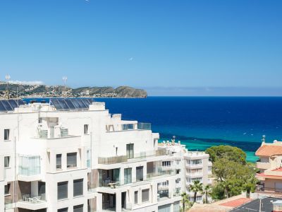 Panoramablick auf moderne weiße Wohngebäude mit Glasbalkonen, Solarpaneelen auf den Dächern und das tiefblaue Mittelmeer bis zur fernen Küstenlinie.