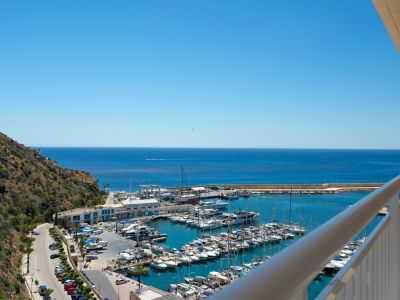 Panoramablick vom Balkon auf einen belebten Yachthafen mit zahlreichen Booten, das tiefblaue Meer und einen grünen Hügel unter klarem Himmel.