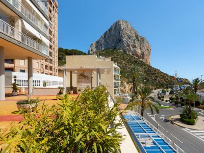 Panoramablick von einer Terrasse mit üppigen Pflanzen im Vordergrund, über die Küstenstraße, den Hafenbereich und den majestätischen Peñón de Ifach.