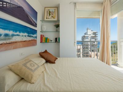 Schlafzimmer mit gemütlichem Bett, maritimem Wandschmuck und Glasregalen. Direkter Zugang zu einem Balkon mit teilweisem Meer- und Stadtblick.