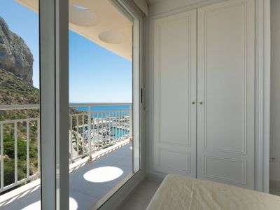 Schlafzimmer mit weißem Einbauschrank und Terrassenzugang. Panoramablick auf den Peñón de Ifach, den Yachthafen und das Mittelmeer unter blauem Himmel.