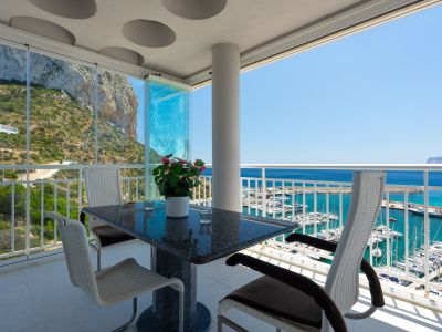Überdachte Terrasse mit Granittisch und modernen Stühlen, ausgestattet mit Glasvorhängen, die einen Panoramablick auf den Peñón de Ifach und den Hafen von Calpe bietet.