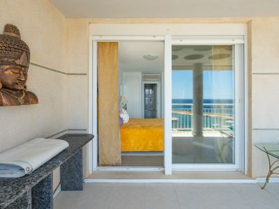 Überdachte Terrasse mit hellen Fliesen und strukturierten Wänden, mit Glasschiebetür zu einem Schlafzimmer, von dem aus Meerblick sichtbar ist. Eine Granitkonsole und Deko sind ebenfalls zu sehen.
