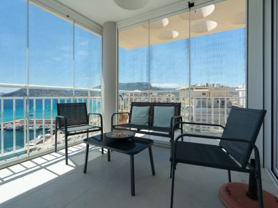 Überdachte Terrasse mit verschiebbaren Glasvorhängen, Outdoor-Möbeln und Panoramablick auf den Hafen von Calpe, das Meer und die Berge. Heller Fliesenboden.