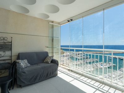 Verglaste Terrasse mit direktem Blick auf den Yachthafen und das Meer. Ausgestattet mit Sofa und schmiedeeisernem Regal, unter einer Decke mit runden Dekorelementen.