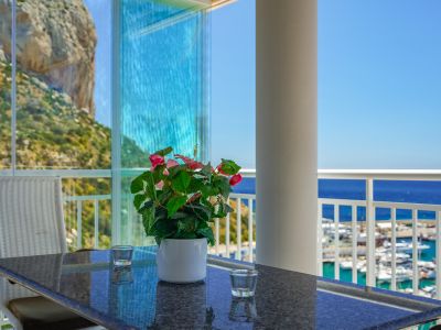 Verglaste Terrasse mit Glasvorhängen, direktem Blick auf den Peñón de Ifach und den Yachthafen von Calpe. Granittisch mit Blumenarrangement.