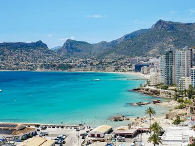 Weitläufiges Küstenpanorama der Bucht von Calpe, mit türkisfarbenem Wasser, Sandstränden, modernen Hochhäusern und fernen Bergen. Der Hafen mit Solarpaneelen ist im Vordergrund.
