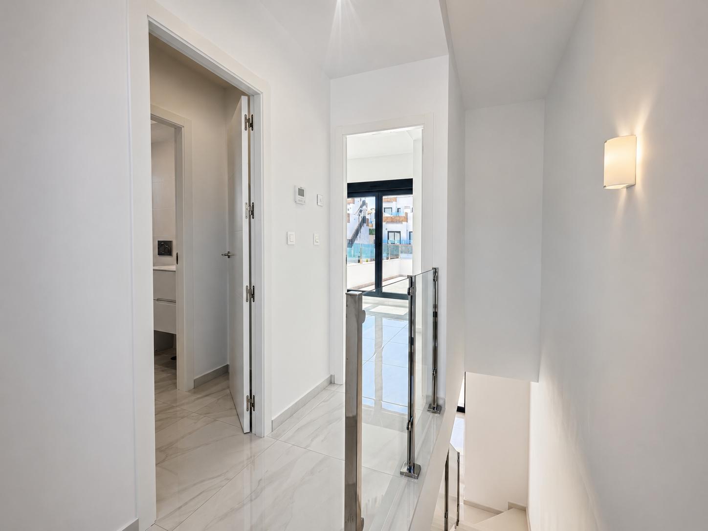 Heller Flur im Obergeschoss mit polierten Marmoroptik-Fliesen und modernem Glas-Stahl-Treppengeländer. Eine Tür führt zum Badezimmer, eine andere zu einem Außenbalkon mit Tageslicht.