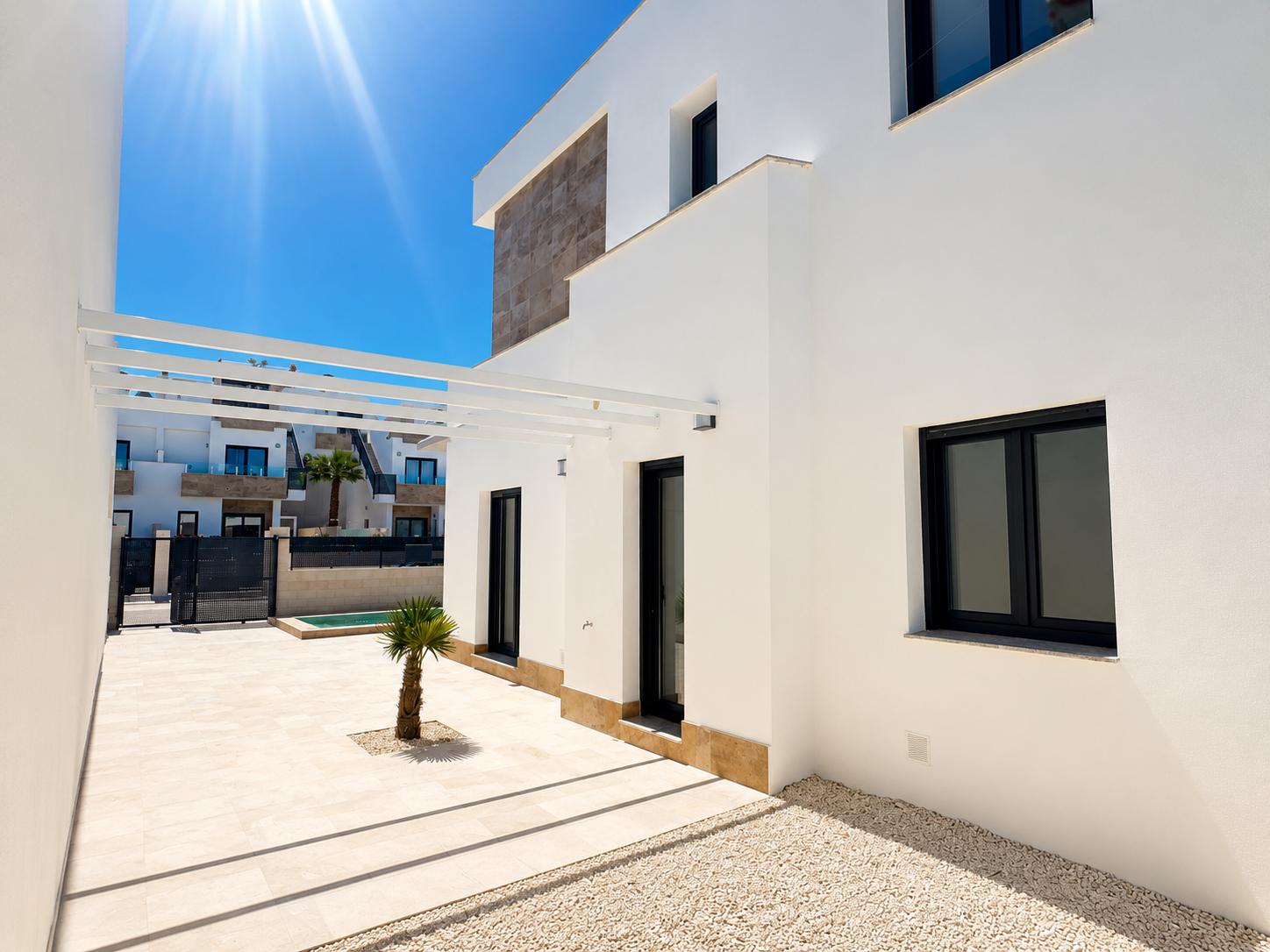 Moderner Villa-Außenbereich mit weißer Fassade und Steinakzenten. Helle Fliesenterrasse, kleine Palme, weiße Pergola und dunkel gerahmte Fenster bei Sonnenschein.