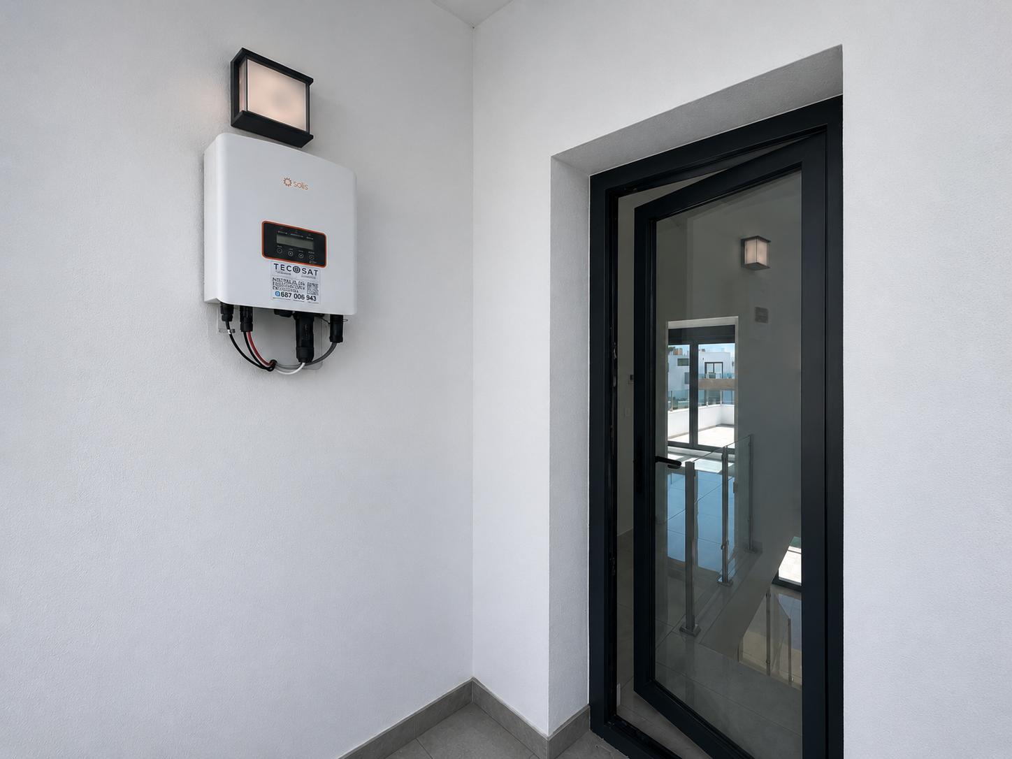 Solis Solarwechselrichter an weißer Wand mit Außenleuchte. Eine halb geöffnete Glastür mit schwarzem Rahmen gibt den Blick auf eine Innentreppe mit Geländer frei.