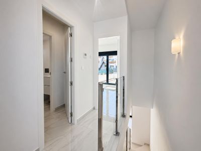 Heller Flur im Obergeschoss mit polierten Marmoroptik-Fliesen und modernem Glas-Stahl-Treppengeländer. Eine Tür führt zum Badezimmer, eine andere zu einem Außenbalkon mit Tageslicht.