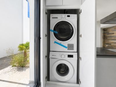 Integrierter Waschbereich mit gestapelter Waschmaschine und Trockner, diskret in weißen Schränken versteckt. Ein Blick auf den Garten und die moderne Küche ist sichtbar.