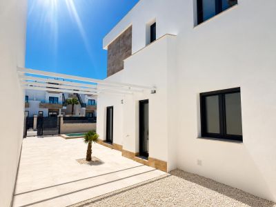 Moderner Villa-Außenbereich mit weißer Fassade und Steinakzenten. Helle Fliesenterrasse, kleine Palme, weiße Pergola und dunkel gerahmte Fenster bei Sonnenschein.
