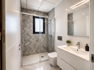Modernes Badezimmer mit Glasduschkabine, grauen geometrischen Fliesen und Fenster. Weißer Waschtisch, LED-beleuchteter Spiegel und Marmoroptik-Boden.