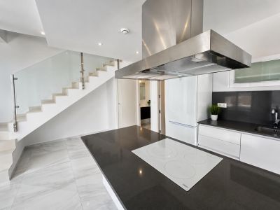 Offene Küche mit dunkler Arbeitsplatteninsel und weißem Induktionskochfeld, daneben eine moderne Treppe mit Glasgeländer und weißen Stufen.