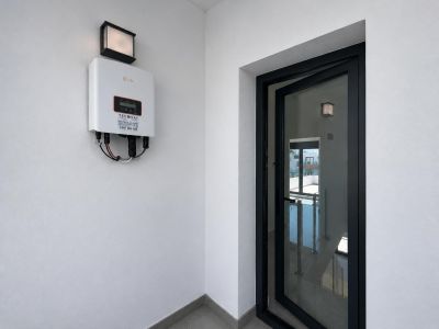 Solis Solarwechselrichter an weißer Wand mit Außenleuchte. Eine halb geöffnete Glastür mit schwarzem Rahmen gibt den Blick auf eine Innentreppe mit Geländer frei.