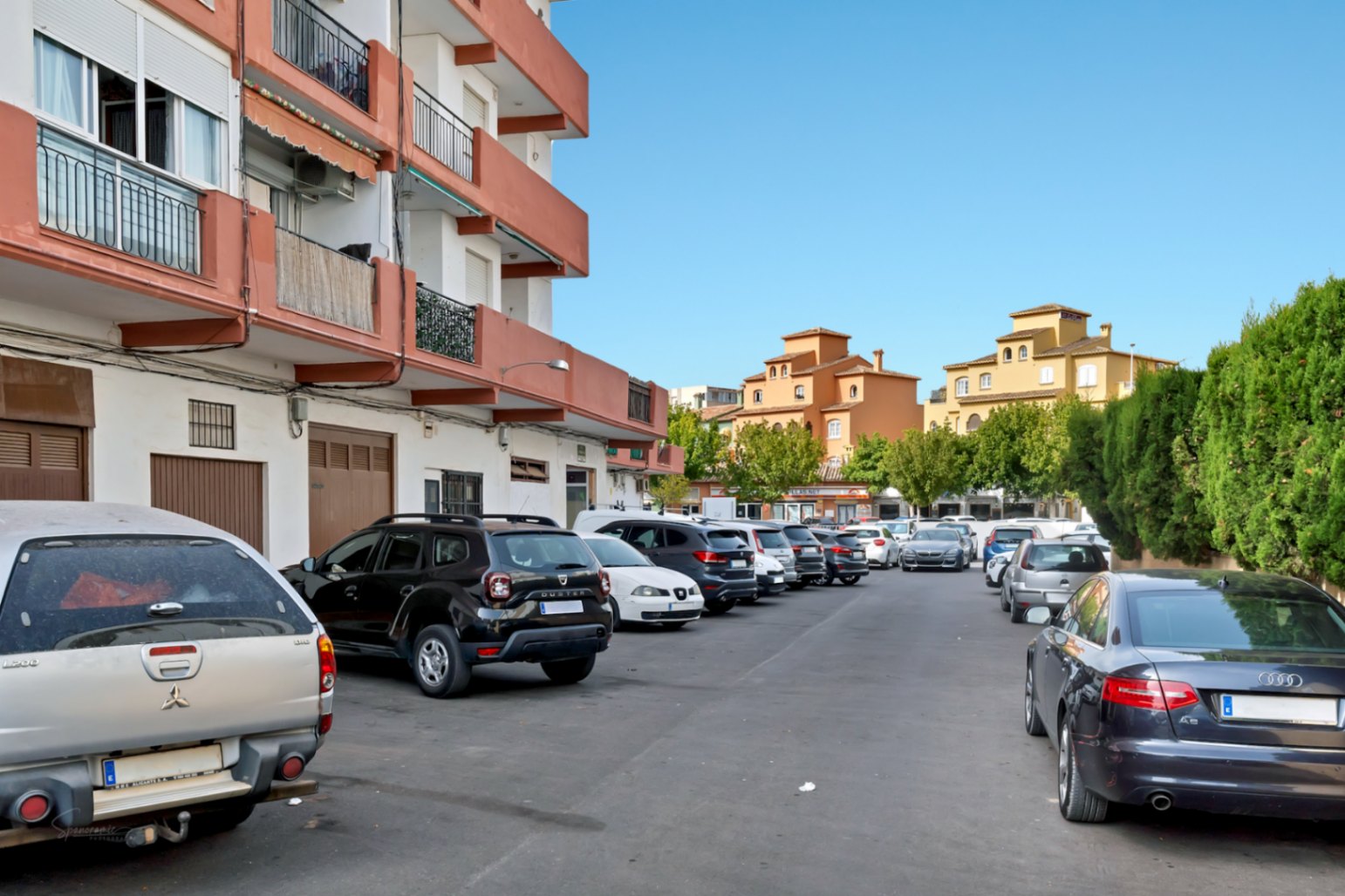 Außenparkplatz mit mehreren Fahrzeugen, gesäumt von einem Wohngebäude mit orangefarbenen Balkonen und traditionellen ockerfarbenen Gebäuden im Hintergrund bei klarem Himmel.