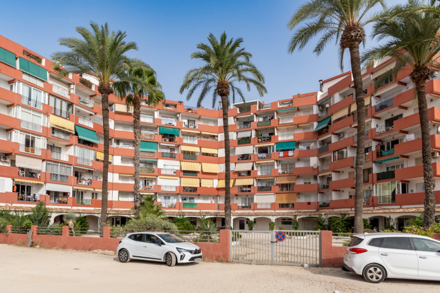 Geschwungenes Apartmentgebäude mit Terrakotta-Fassade und Balkonen mit verschiedenen Markisen. Hohe Palmen im Vordergrund, davor ein Außenparkplatz mit Tor.