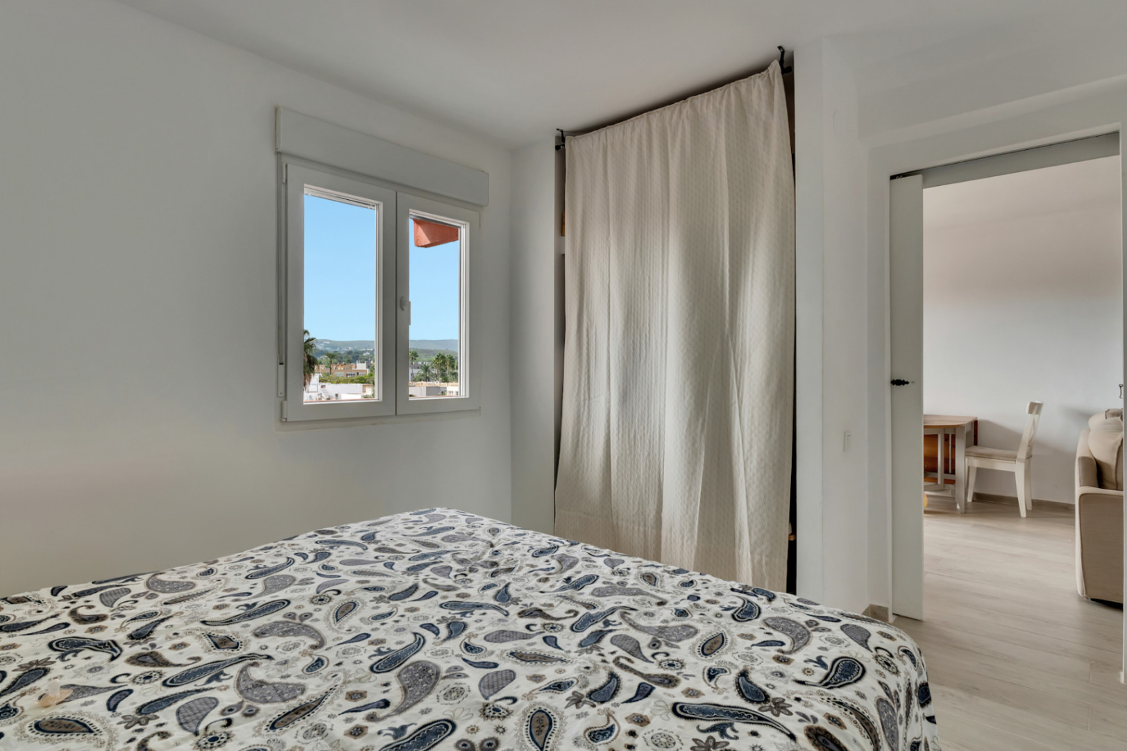 Helles Schlafzimmer mit gemustertem Bettbezug, Fenster mit Stadtblick und einem dezenten, von einem hellen Vorhang verdeckten Kleiderschrank, mit Zugang zu einem Nebenraum.