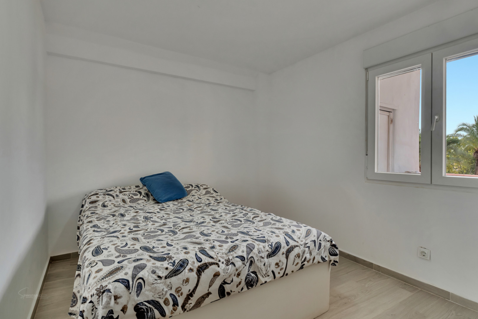 Helles Schlafzimmer mit weißen Wänden und hellem Laminatboden. Ein Bett mit Paisley-Bettdecke und blauem Kissen steht an einem Fenster, das Tageslicht und einen Blick auf Palmen bietet.