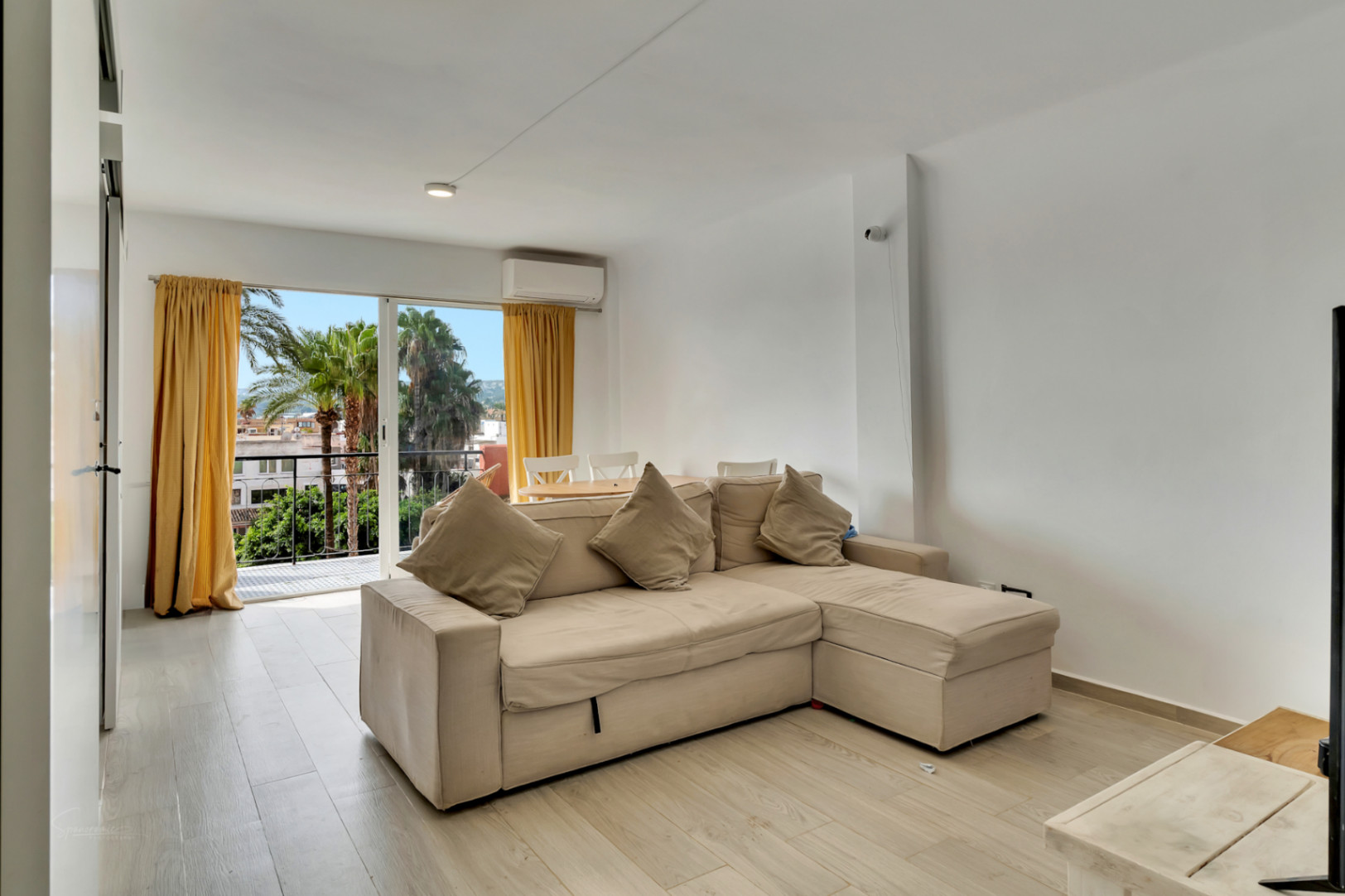 Helles Wohnzimmer mit beigem Ecksofa, hellem Holzfußboden und Balkonzugang mit Palmenblick und gelben Vorhängen.