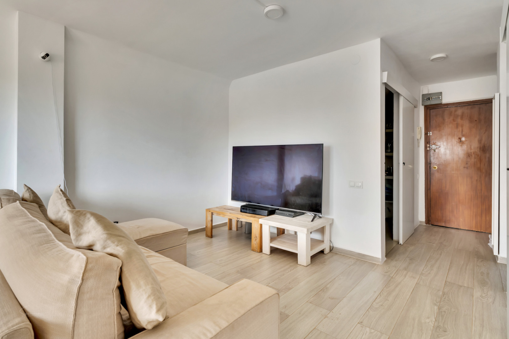 Helles Wohnzimmer mit beigem Ecksofa und TV auf hellen Holztischen. Der helle Laminatboden führt zur dunklen Holzeingangstür und einem offenen Regalbereich.