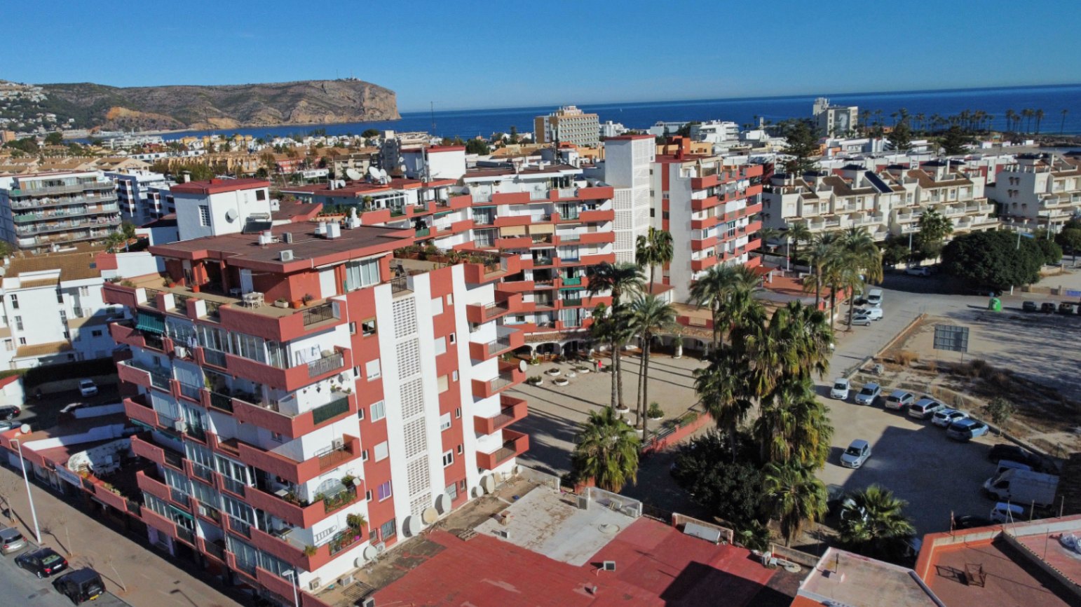 Luftaufnahme des Wohnkomplexes mit weißen Fassaden, rot-orangen Balkonen, Palmen und dem Mittelmeer im Hintergrund.