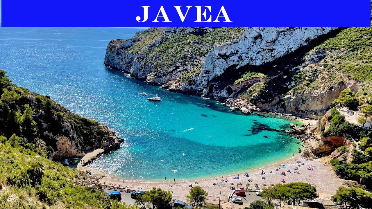 Luftaufnahme einer Bucht in Jávea mit klarem türkisfarbenem Wasser, einem Kiesstrand mit Badegästen und felsigen Klippen, die von mediterraner Vegetation bewachsen sind.