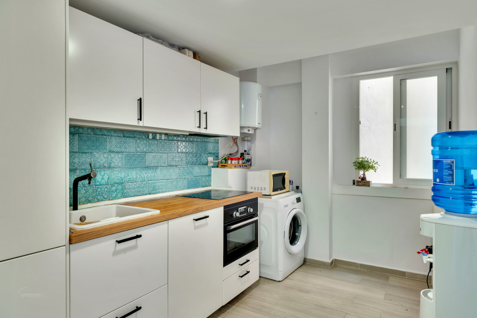 Moderne Küche mit weißen Schränken, heller Holztheke und einem lebhaften türkisfarbenen Fliesenspiegel. Mit integrierten Geräten und einem Hauswirtschaftsbereich mit Waschmaschine.