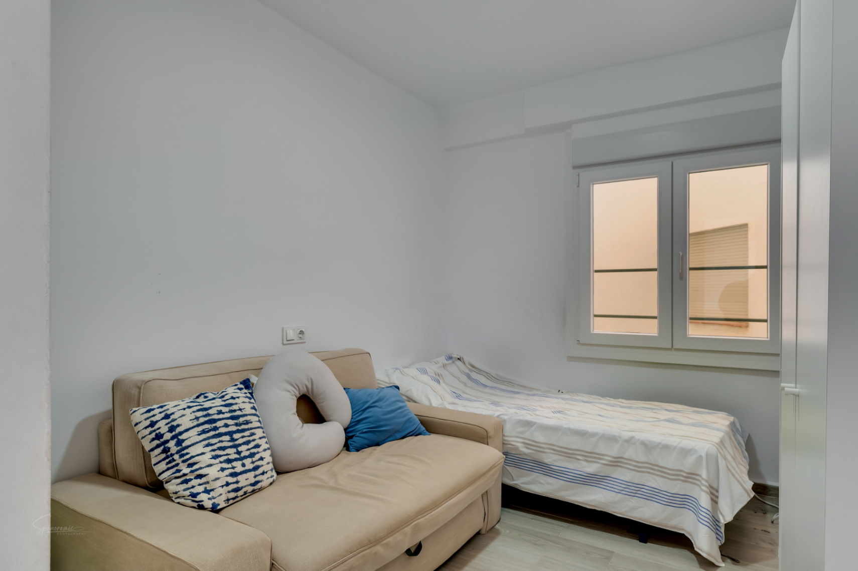 Nebenzimmer mit beigefarbenem Schlafsofa und Einzelbett, weißen Wänden und Fenster mit diffusem Tageslicht.