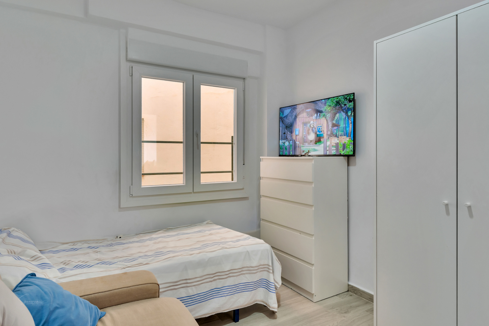 Nebenzimmer mit Einzelbett, weißem Einbauschrank und Kommode mit Wandfernseher. Das Doppelfenster sorgt für natürliches Licht.