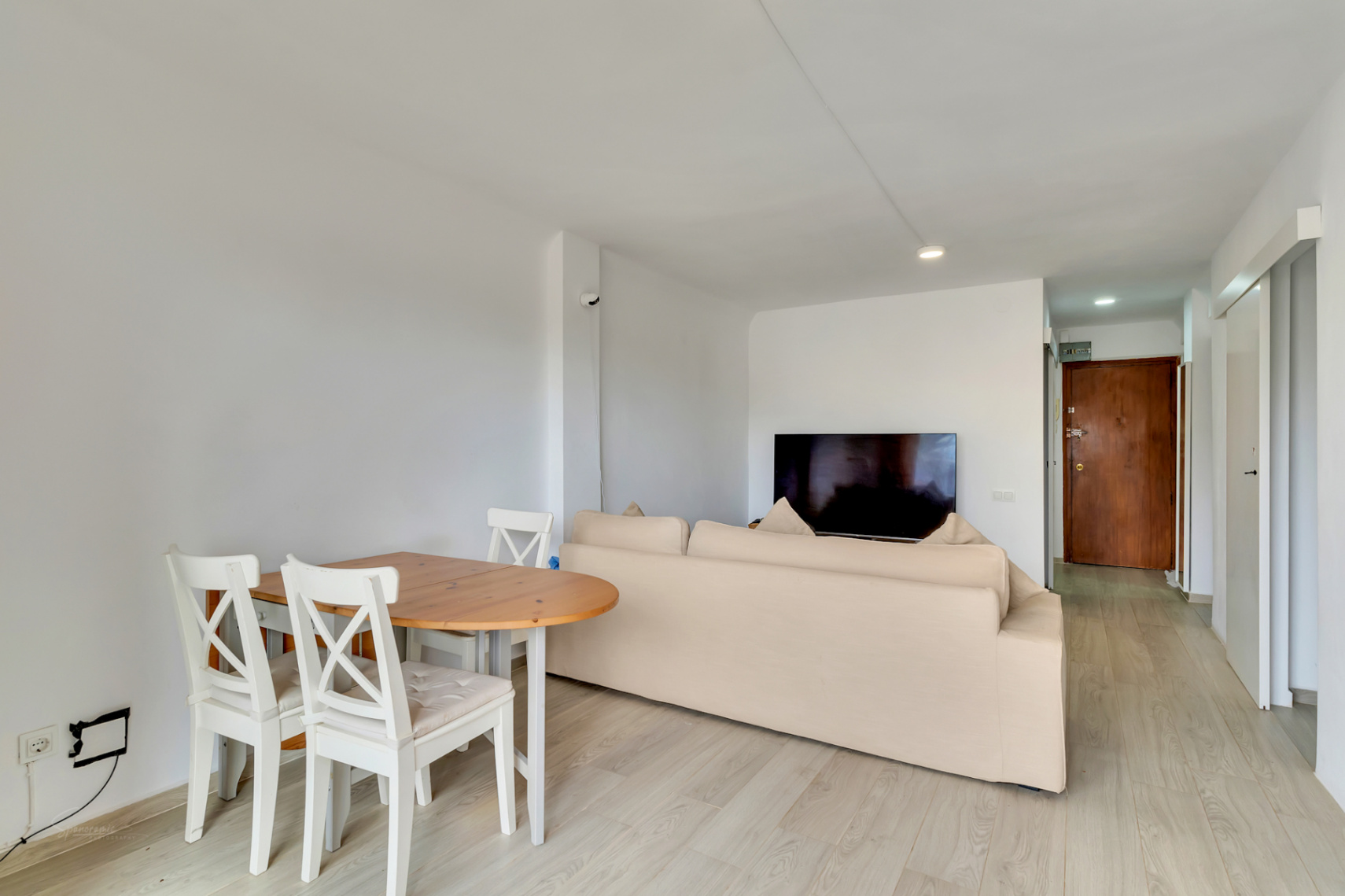 Offener Wohn- und Essbereich mit hellem Holztisch, weißen Stühlen und beigem Sofa. Ein Wandfernseher und heller Laminatboden führen zu einer dunklen Holzeingangstür.
