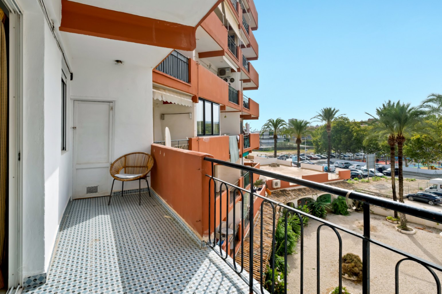 Sonniger Balkon mit blau-weißem geometrischem Fliesenboden, ausgestattet mit einem Korbstuhl und Abstellraumtür. Blick auf Palmen und den Wohnparkplatz.