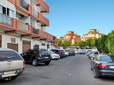 Außenparkplatz mit mehreren Fahrzeugen, gesäumt von einem Wohngebäude mit orangefarbenen Balkonen und traditionellen ockerfarbenen Gebäuden im Hintergrund bei klarem Himmel.