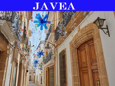 Enge Gasse im historischen Zentrum von Javea, mit traditionellen Steintüren, schmiedeeisernen Balkonen und festlichen Hängedekorationen mit blauen Sternformen.