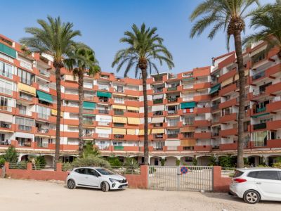 Geschwungenes Apartmentgebäude mit Terrakotta-Fassade und Balkonen mit verschiedenen Markisen. Hohe Palmen im Vordergrund, davor ein Außenparkplatz mit Tor.