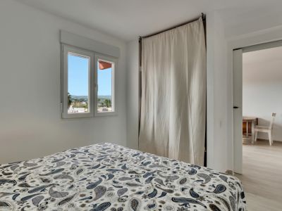 Helles Schlafzimmer mit gemustertem Bettbezug, Fenster mit Stadtblick und einem dezenten, von einem hellen Vorhang verdeckten Kleiderschrank, mit Zugang zu einem Nebenraum.
