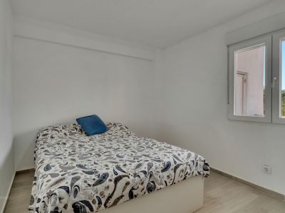 Helles Schlafzimmer mit weißen Wänden und hellem Laminatboden. Ein Bett mit Paisley-Bettdecke und blauem Kissen steht an einem Fenster, das Tageslicht und einen Blick auf Palmen bietet.