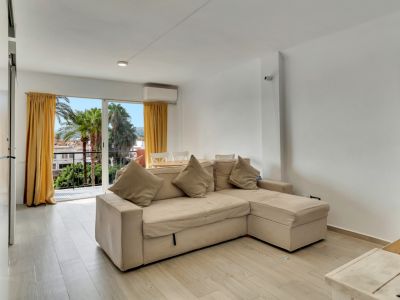 Helles Wohnzimmer mit beigem Ecksofa, hellem Holzfußboden und Balkonzugang mit Palmenblick und gelben Vorhängen.